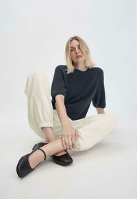 Neulottu tummansininen lyhythihainen villapaita, kerma-valkoiset elastiset housut ja mustat nilkkavöillä varustetut balettikengät. Malli poseeraa istuen, kädet jalkojen ympärillä.