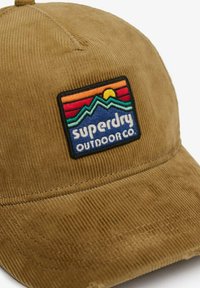 Braune Corduroy-Mütze mit einem bunten gestickten Patch, der Berge, die Sonne und den Text "Superdry Outdoor Co." auf der Vorderseite zeigt.