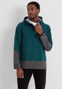 Man wearing a teal and gray hooded zip-up jacket, adjusting the zipper with his right hand, standing against a plain white background.