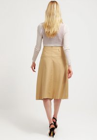 Žena s dlouhými blond vlasy, oblečená v béžové midi sukni, světle pleteném krátkém svetru a černých lodičkách na vysokém podpatku, stojící zády.