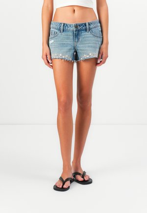 Persona che indossa pantaloni corti di jeans azzurri con ricami floreali e infradito nere, in piedi contro uno sfondo bianco semplice.