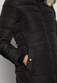 Černá prošívaná bunda s horizontálními švy, zipovým zapínáním a bočními kapsami. Obsahuje žebrované manžety a kapuci vystlanou kožešinou.