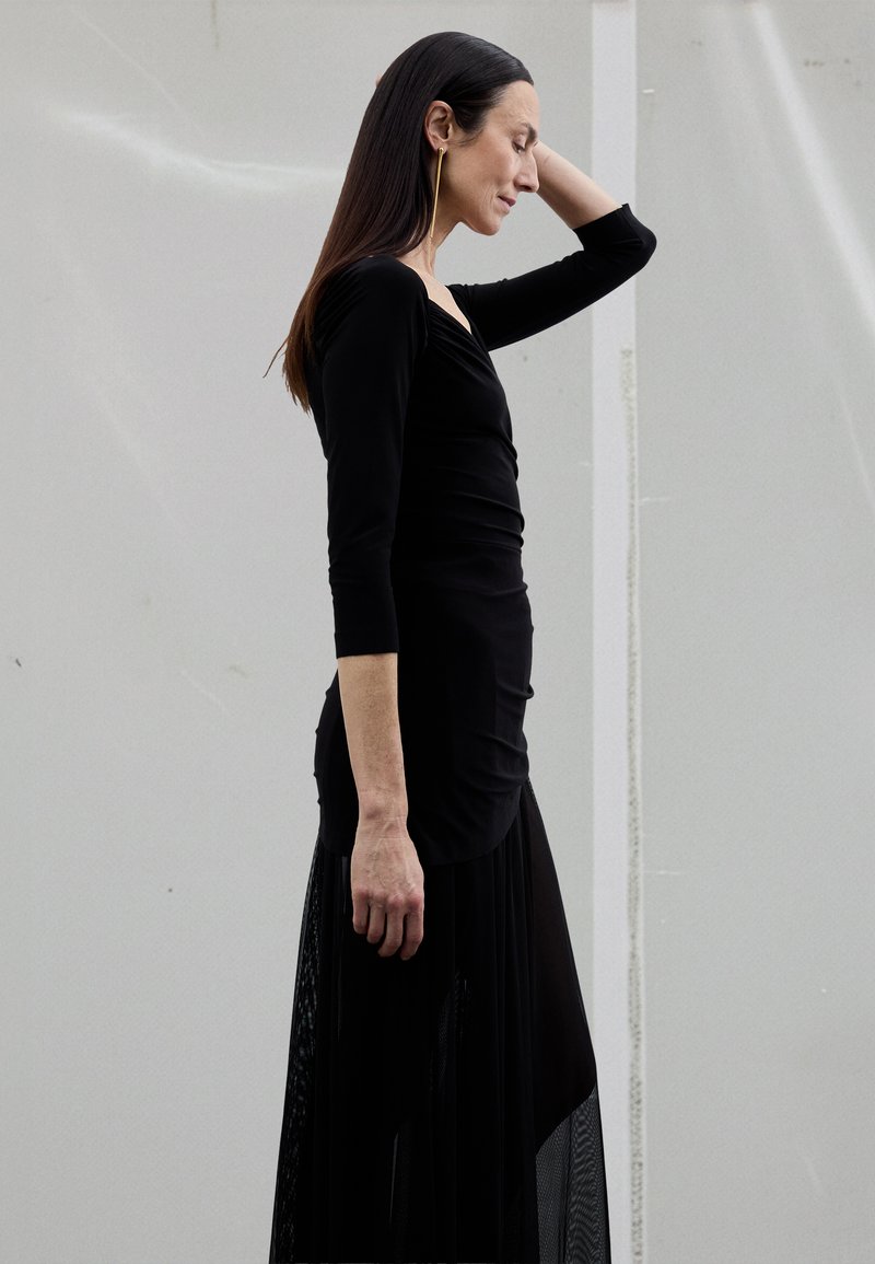 Rochie neagră cu partea de sus mulată și mâneci lungi, cu o fustă fluidă și un strat exterior transparent. Textură netedă, detalii minimale.