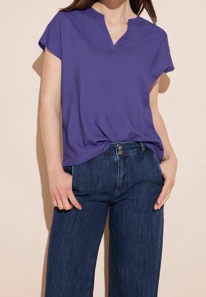 Femme portant un chemisier violet à manches courtes avec un col en V et un jean bleu foncé taille haute, les mains dans les poches, debout devant un fond beige.