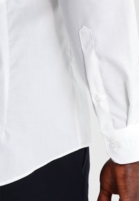 Vit långärmad skjorta med en mjuk textur, spetsig manschett och två knappar. Nederkanten är lätt rundad med sidoslitsar för komfort.