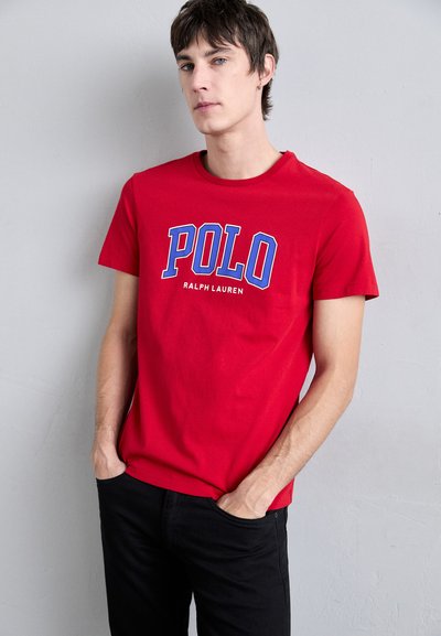 Piros pamut póló, kék és fehér "POLO" logóval, kerek nyakkivágással és rövid ujjakkal, fekete farmernadrággal párosítva, texturált háttérrel.