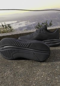 Paire de chaussures de sport noires posées sur un sol en béton avec une ville et un paysage flous en arrière-plan à la lumière du jour.