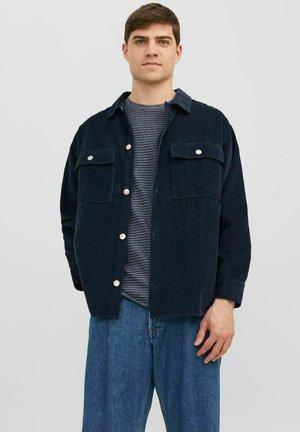 Veste chemise en velours côtelé bleu marine avec deux poches avant, fermeture à boutons et coupe décontractée, associée à un t-shirt rayé et un jean bleu.