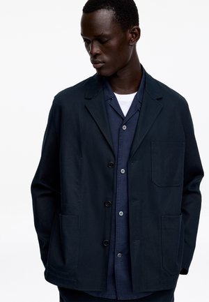 Mężczyzna w warstwowej ciemnogranatowej kurtce na koszulę z guzikami i białą podkoszulką, patrzący w dół na jednolitym białym tle.