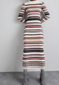 Randig stickad klänning med långa ärmar, ribbad textur och färger som inkluderar vitt, rosa, svart och beige. Längd till mitten av vaden.