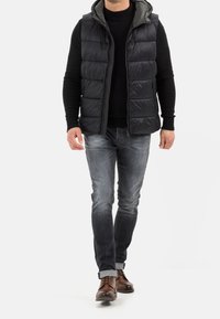 Schwarze Steppweste mit Kapuze, mit Reißverschluss vorne und Seitentaschen, über einem schwarzen Pullover getragen. Kombiniert mit grauen Jeans und braunen Lederschuhen.