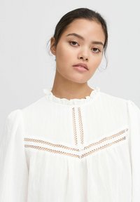 Witte blouse met kraag van ruffles, lange poffende mouwen en kantdetails aan de voorkant. Zachte, gestructureerde stof met subtiele doorschijnendheid.