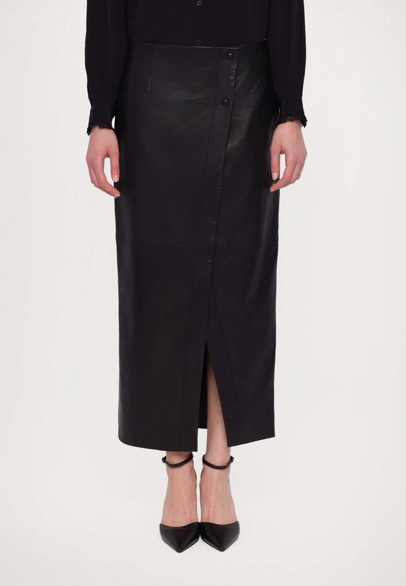 Falda midi de cuero negro con diseño envolvente, botones laterales y una abertura en el frente. Combinada con tacones negros puntiagudos y delicadas correas en el tobillo.