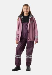 Fille portant une salopette imperméable violette avec des bandes réfléchissantes, une veste de pluie rose, un t-shirt noir et des bottes noires, debout et souriante.