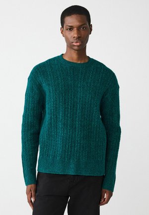 Türkiser Strickpullover mit geripptem Muster, rundem Halsausschnitt und langen Ärmeln, kombiniert mit schwarzen Hosen. Weiches, lässiges Material.