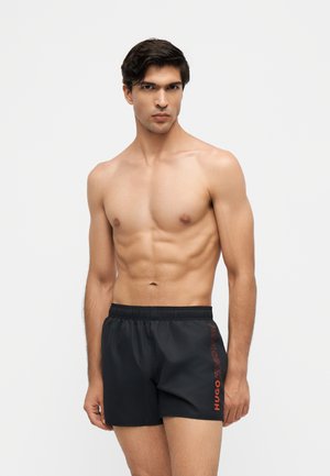 Man zonder shirt met kort donker haar, die zwarte zwemshorts draagt met een rood Hugo-logo op het linkerbeen, tegen een effen achtergrond.