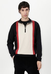 Svart og kremfarget sweatshirt med kvart glidelås, geometrisk mønster på kremfarget panel og røde side striper. Myk stoff og avslappet passform.