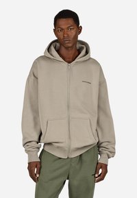 Šviesiai rudas gobtuvu užsegamas džemperis su raišteliu, kišenėmis kaip ant kengūros ir briaunuotais rankogaliais. Dėvimas su atitinkančiomis alyvuogių žalios spalvos sportinėmis kelnėmis.