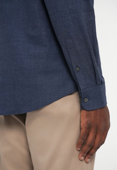 Camisa azul-marinho feita de tecido texturizado, com botões pretos nos punhos, com uma barra curva e combinada com calças bege.