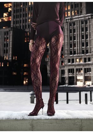 Osoba nosící burgundské krajkové punčochy a ladící otevřené lodičky na podpatku stojící na sněhem pokrytém okraji v nočním městském prostředí.