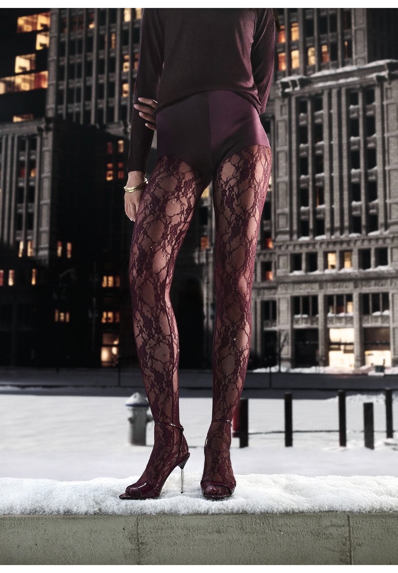 Persona che indossa calze di pizzo bordeaux e tacchi aperti abbinati, in piedi su un cornicione coperto di neve in un ambiente urbano notturno.