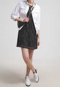 Robe noire àpois blancs, superposée sous une veste en jean blanche. Le mannequin porte un sac bandoulière noir et des bottines blanches à cheville.