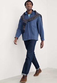 Blå långärmad skjorta med halvdragkedja, mörka denimjeans och bruna ankelstövlar. En mönstrad scarf hänger över skjortans axlar.