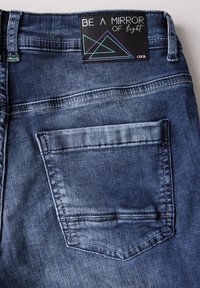 Jeans de denim en azul oscuro con un acabado desgastado. Cuenta con un bolsillo trasero y una etiqueta negra con un diseño geométrico y texto.