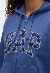 Personne portant un sweat à capuche bleu zippé avec des lettres à motif floral formant le mot "GAP" sur la poitrine et des cordons à la capuche.