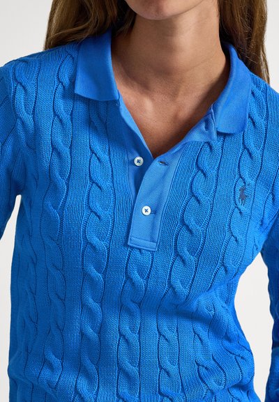 Artimos planas žmogaus, dėvinčio ryškiai mėlyną megztinį su pintu raštu, polo apykakle ir dviem užsegamomis sagomis prie kaklo.