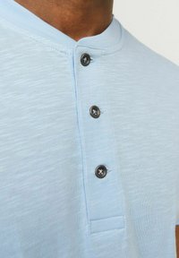 Camisa de botones azul claro con una tela texturizada, que presenta tres botones oscuros y un cuello redondo. Diseño casual, vista en primer plano.