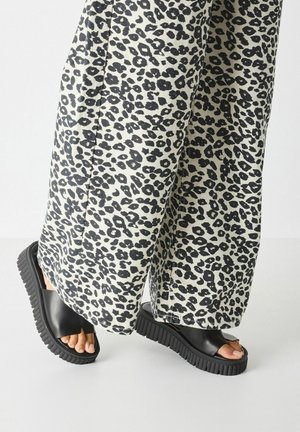 Sandalias de tiras negras con suela gruesa y texturizada, usadas con pantalones de pierna ancha que presentan un patrón de estampado de leopardo en blanco y negro.