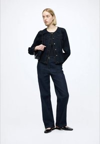 Tumša džinsa jaka ar zelta pogām un kabatām, kombinēta ar platām tumšām džinsu biksēm. Melna somiņa un apavi papildina tērpu.