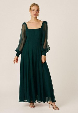 Mujer con un vestido largo verde oscuro con mangas abullonadas transparentes y escote cuadrado, combinado con sandalias doradas de tacón alto sobre un fondo sencillo.