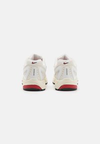 Baltās sporta kurpes ar sieta augšdaļu, pelēkajiem akcentiem un sarkano papēža detaļu. Iekļauj redzamu gaisa amortizācijas vienību un strukturētu zoli.