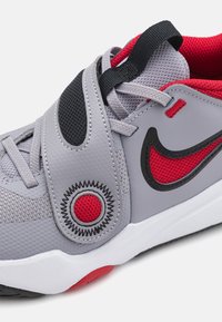 Zapatilla deportiva gris con un upper texturizado, acentos rojos, una correa negra y un distintivo elemento gráfico circular en el lado.