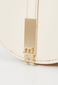 Primo piano dell'angolo di una borsa in pelle crema con cuciture ordinate e una chiusura rettangolare in oro che presenta le lettere "T" e "H" intrecciate.