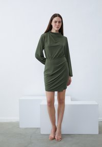 Grünes, langärmeliges Kleid mit drapiertem Design und elastischem Bund. Glattes Gewebe und minimalistische Details, kombiniert mit neutralen Riemchen-Heels.
