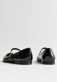 Svart lackläderballetskor med rem och spänne, rundad tå och platt sula. Slät, glänsande yta med minimalistisk design.