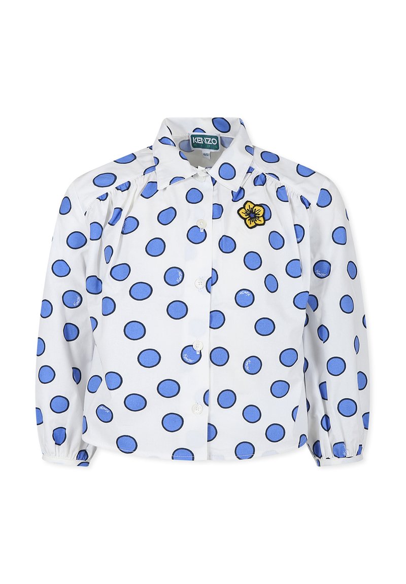 Chemise blanche à manches longues avec des pois bleus, fermeture à boutons, et une broderie de fleur jaune sur la poitrine gauche. Présente des épaules froncées.