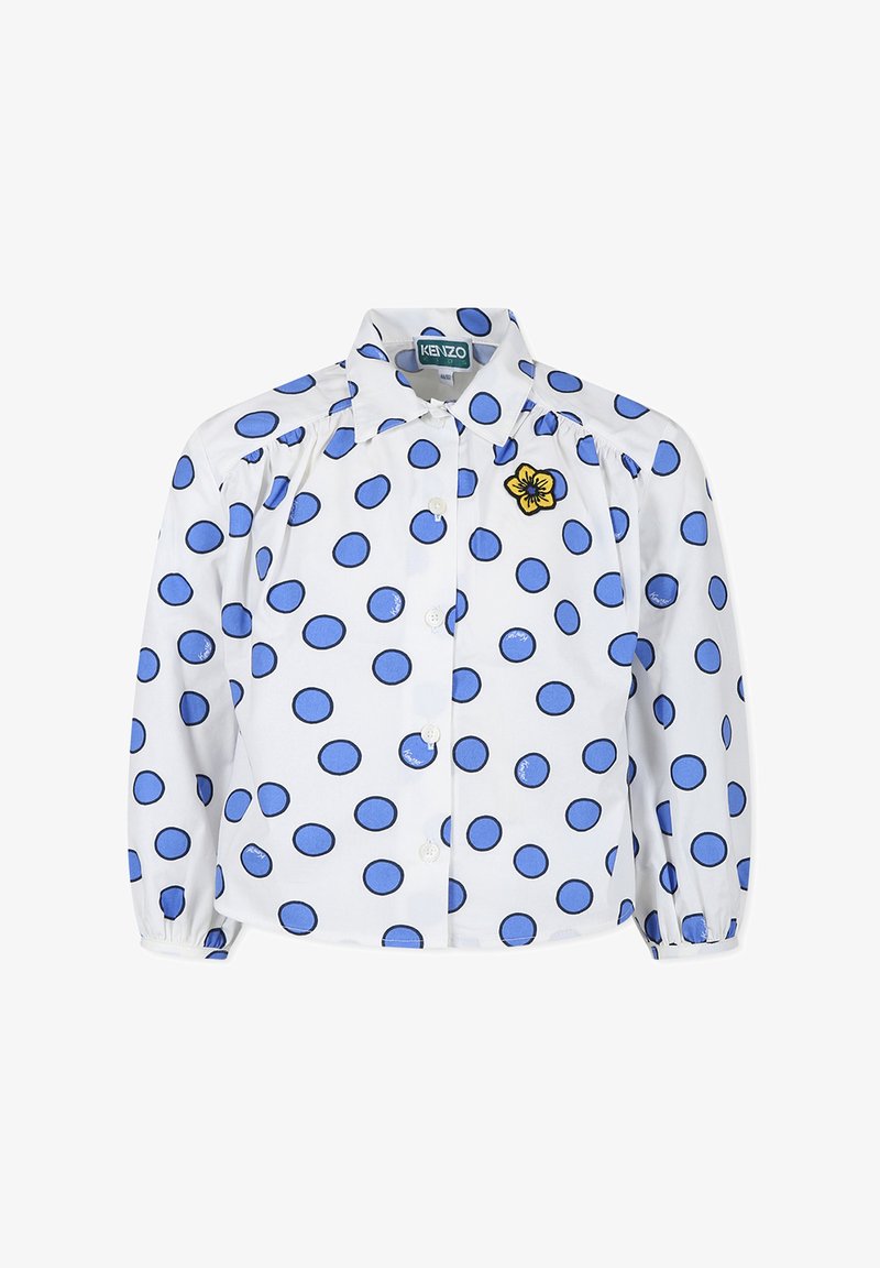 Chemise blanche à manches longues avec des pois bleus, fermeture à boutons, et une broderie de fleur jaune sur la poitrine gauche. Présente des épaules froncées.