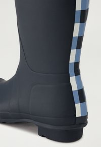 Bota de goma negra con un patrón de cuadros en azul claro y blanco a lo largo del lado. Textura lisa con una suela texturizada para mayor agarre.