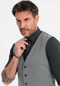 Mann mit kurzen dunklen Haaren und Bart richtet seine marineblaue Krawatte, trägt ein schwarzes Hemd, eine graue Weste und Armbänder, mit sichtbarem tätowiertem Unterarm.