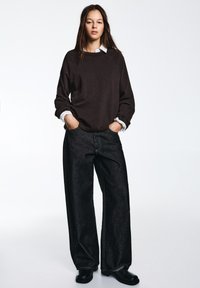 Maglione marrone scuro, camicia bianca con colletto sotto, pantaloni larghi in denim nero e scarpe nere. Presenta scollo a coste e tasche laterali.