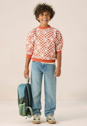 Orange-weiß kariertes Sweatshirt mit Smiley-Gesichtern, kombiniert mit hellblauen Jeans. Die Person hält einen türkisfarbenen Rucksack.