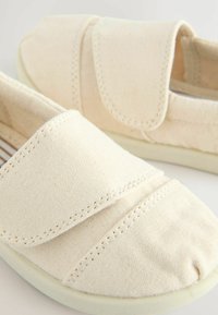 Hellbeige Leinenschlüpfschuhe mit runder Zehenpartie, ausgestattet mit einem Klettverschluss und detaillierter Nahtführung im oberen Bereich. Weiche, flexible Sohle.