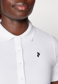 Weißes Poloshirt aus strukturiertem Stoff mit klassischem Kragen, drei Knöpfen und einem kleinen schwarzen gestickten Logo auf der linken Brust.