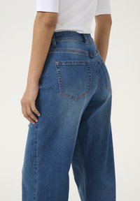 Jeans en denim bleu taille haute à jambes larges avec un lavage délavé, dotés de deux poches arrière et de détails de couture visibles.