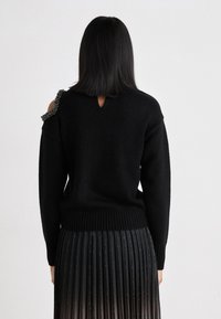 Μαύρο πουλόβερ με ραμμένο στρίφωμα και ώμους, που παρουσιάζει περίπλοκες εκφορές με χάντρες, λεπτομέρεια ανοίγματος στην πλάτη, συνδυασμένο με πτυχωτή μαύρη φούστα.