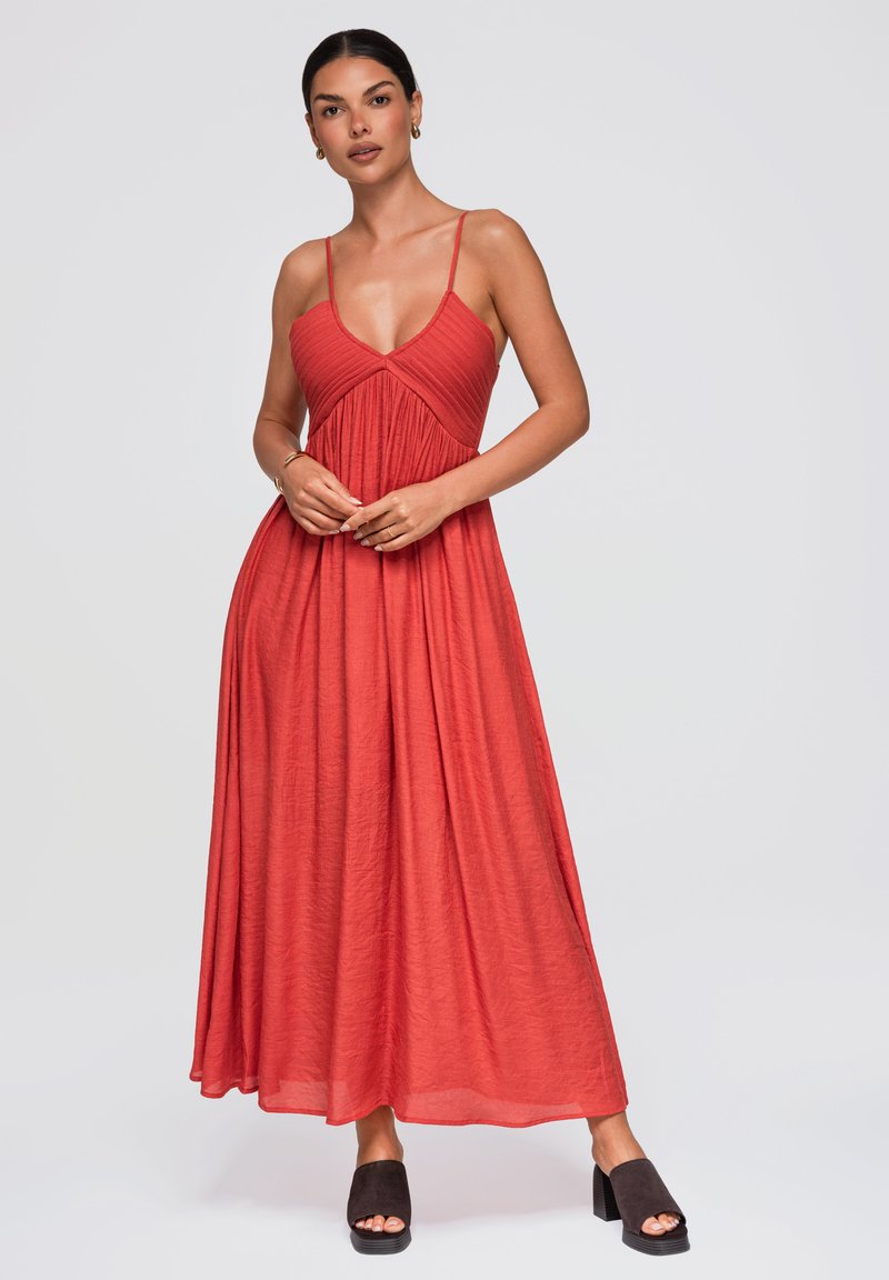 Femme portant une robe longue rouge sans manches avec de fines bretelles, debout les mains jointes et chaussant des sandales noires à talons blocs ouvertes à l'avant.